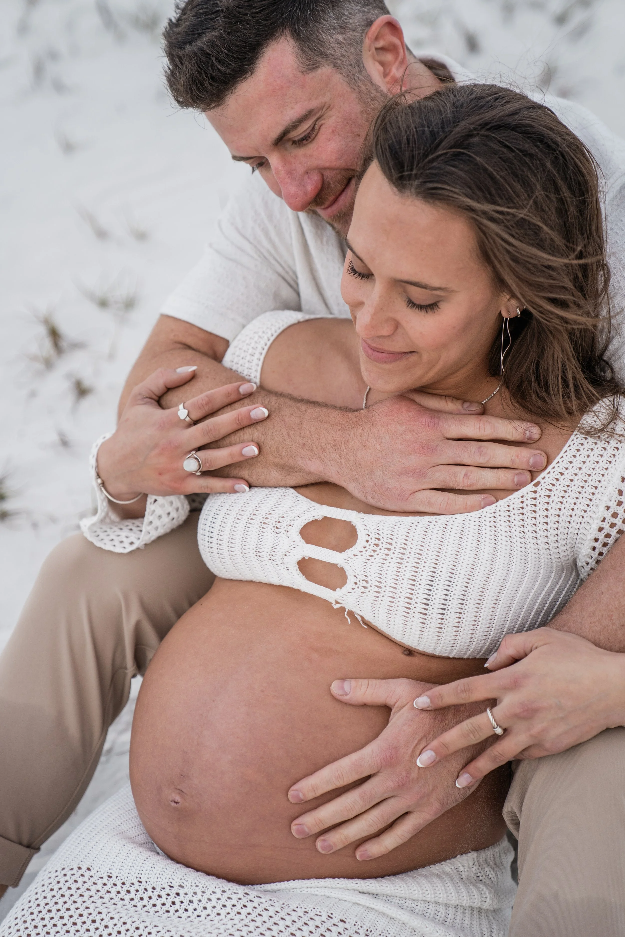 A man and a pregnant woman sitting on white sand, embracing and smiling with closed eyes. The woman is wearing a white crochet top and jewelry, and the man is holding her belly.