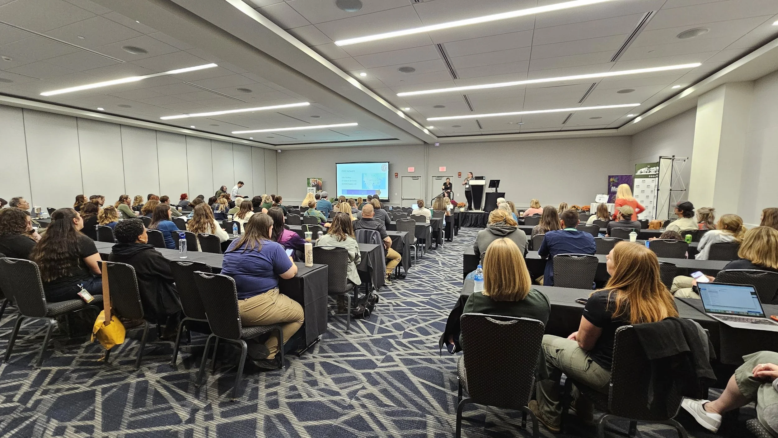 A photo of a full conference room, taken from the back of the room. A presenter stands at the front next to a projector screen