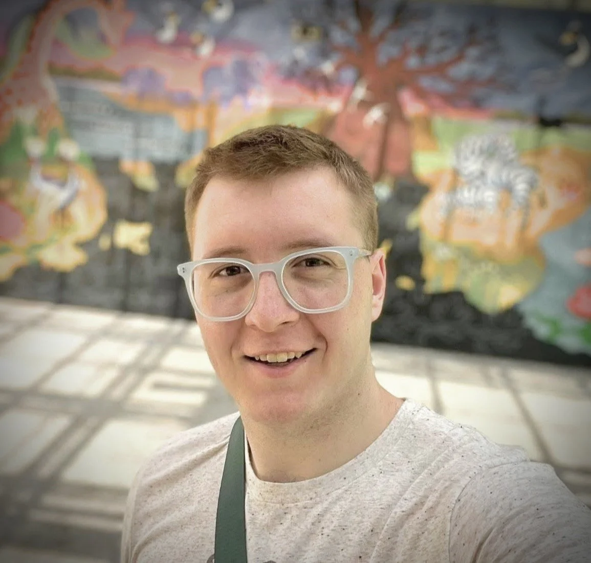 A headshot of Konnor Roberts, a young white man with sandy hair and white-rimmed glasses. He is taking a selfie and smiling.