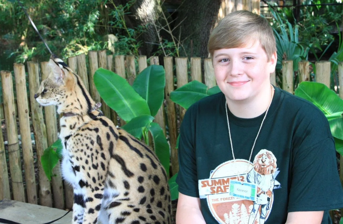 A photo of a young Konnor with an African serval. He is sitting next to the cat in a posed photo, looking into the camera, wearing a shirt that says "Summer Safari"