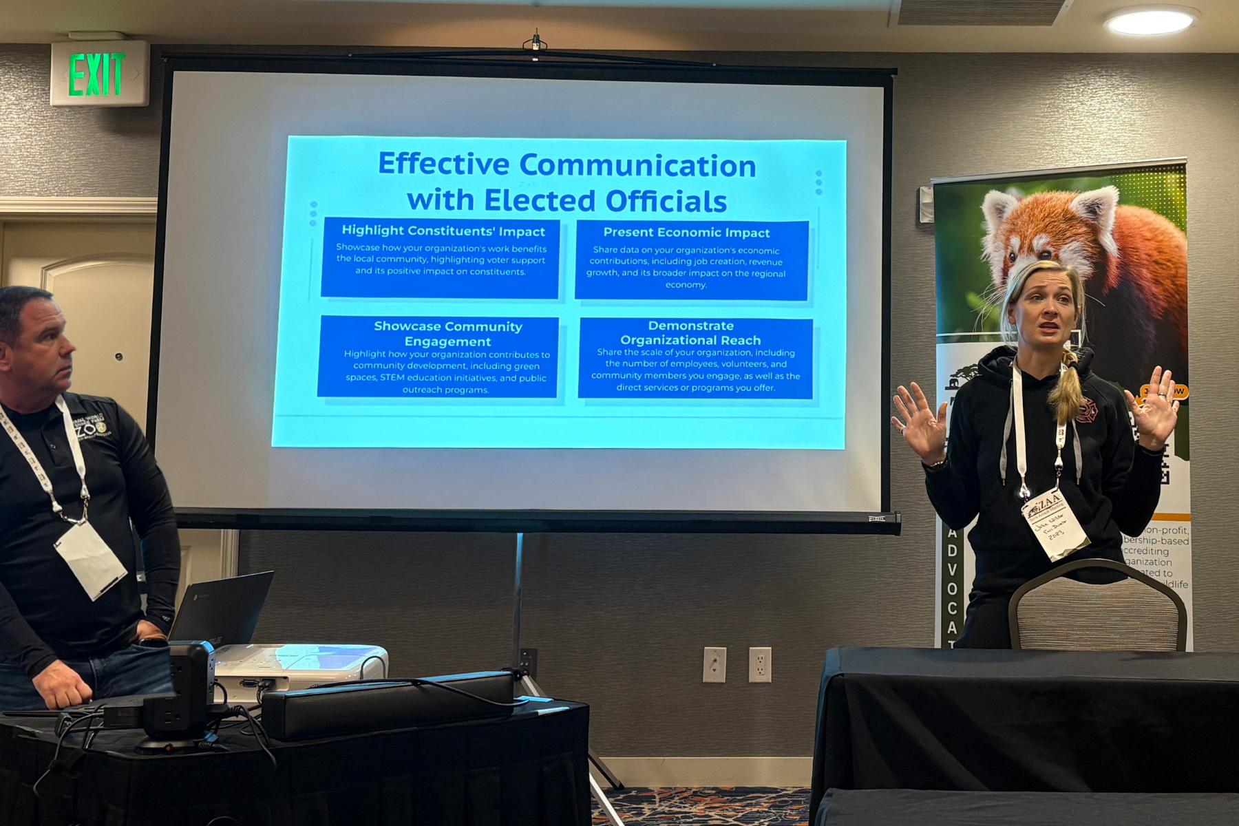 A photo of a blonde woman speaking while standing next to a screen. The screen has a slide from a presentation on it