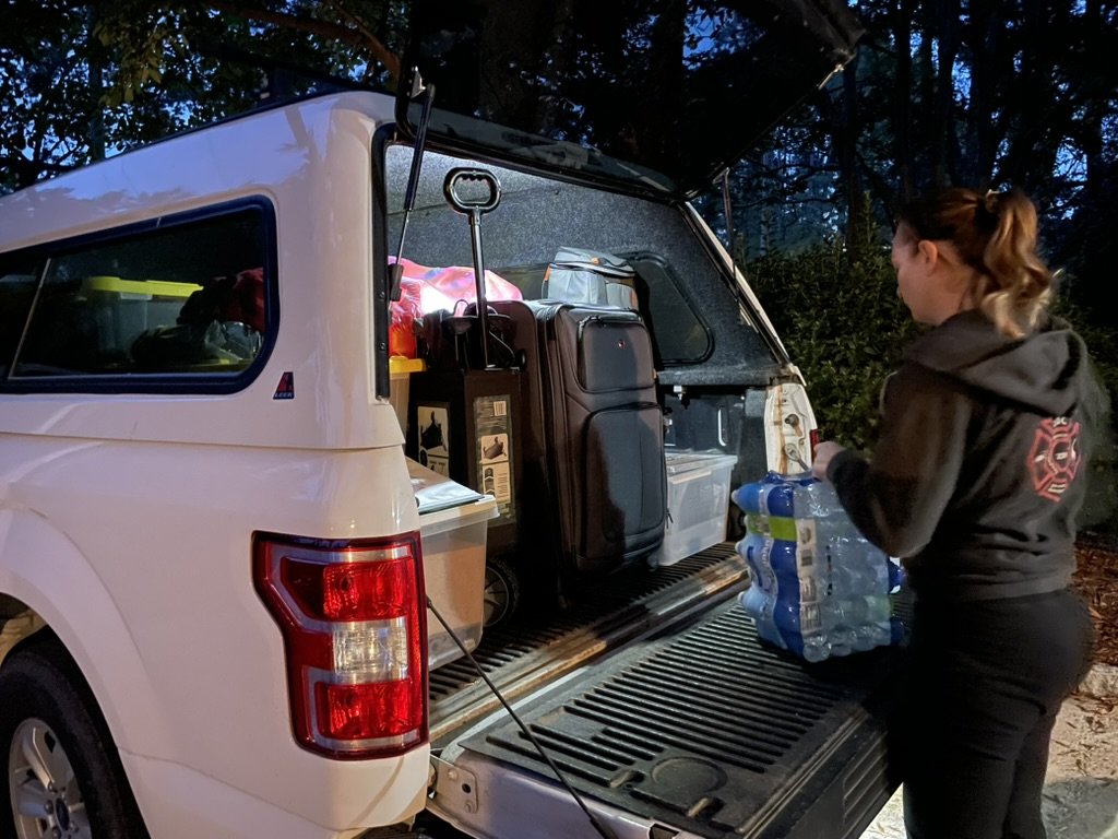 A woman wearing a hoodie with a ZDR3 logo is loading water bottles into the back of a truck with a camper shell packed with supplies and luggage.