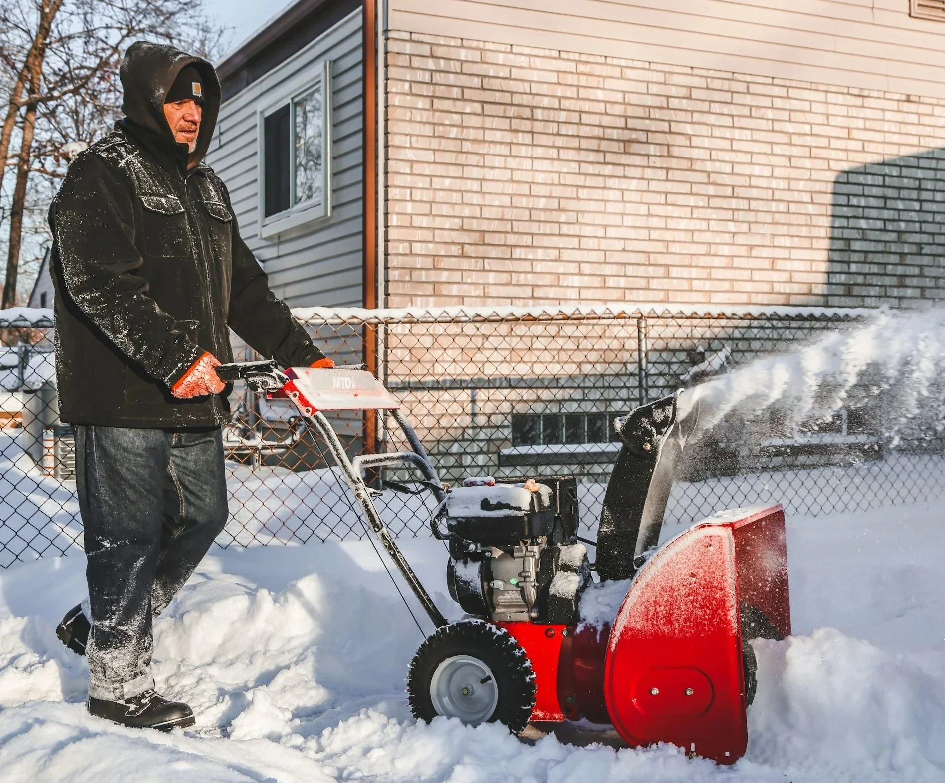 A man in winter outerwear is walking behind a red snow blower, blowing snow