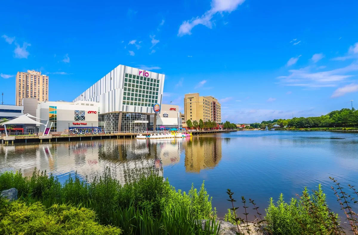 City skyline with modern buildings and a river in the foreground on a clear, sunny day.