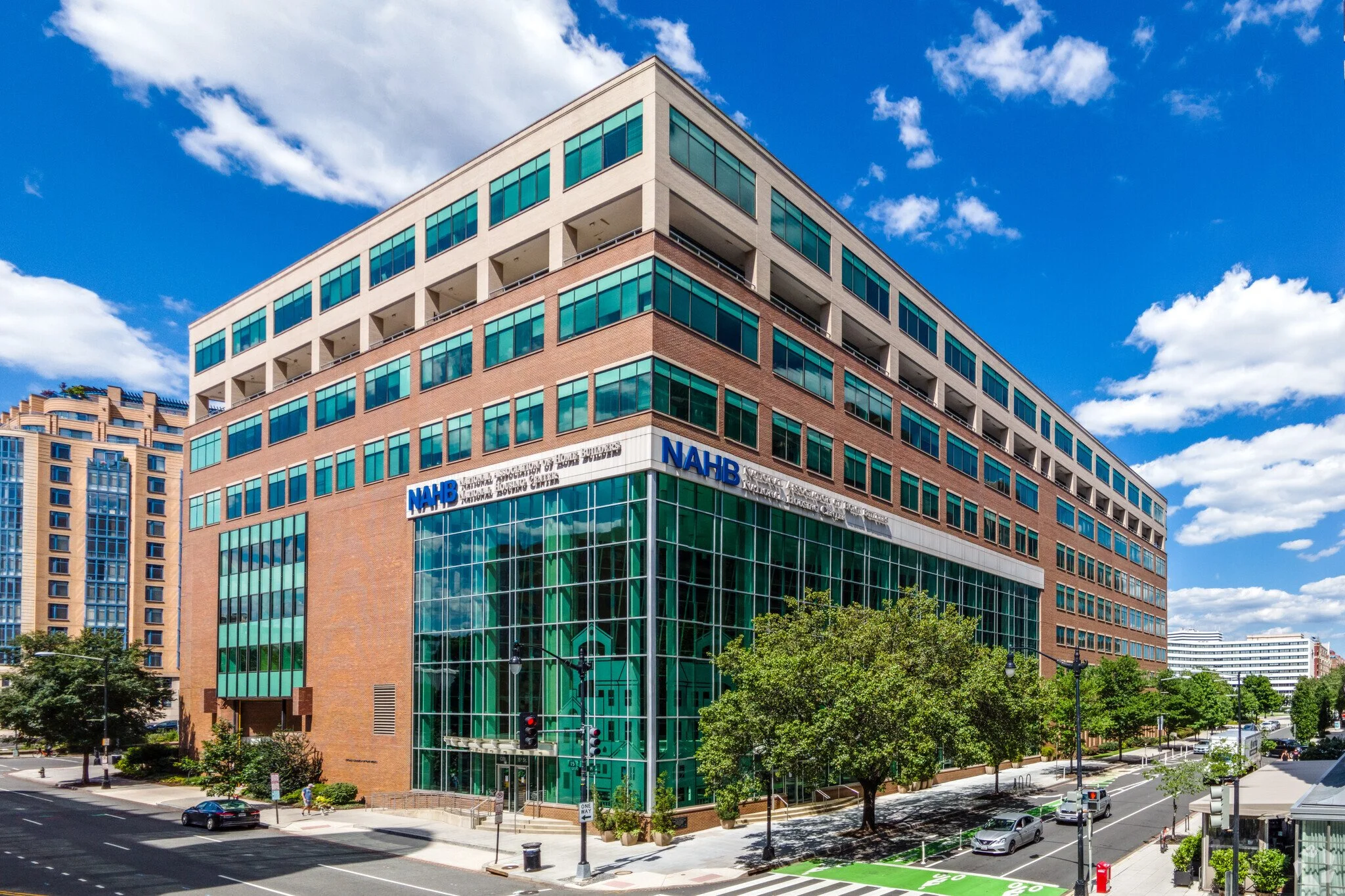 Large modern office building with glass windows and brick facade, located on a city street with trees, cars, and traffic lights under a partly cloudy sky.