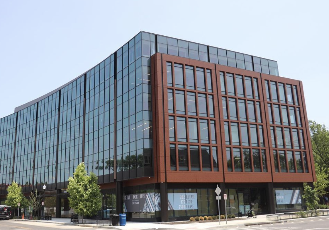 A modern multi-story office building with a glass facade and red brick accents, surrounded by small trees and a sidewalk.