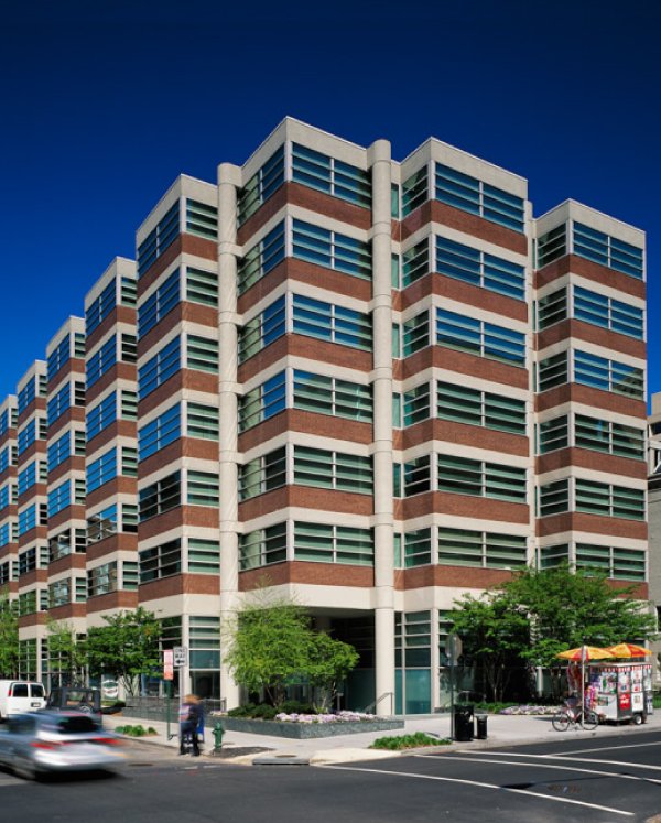 Modern multi-story office building with glass windows and brick accents, situated on a city street with trees, pedestrians, and a food cart.
