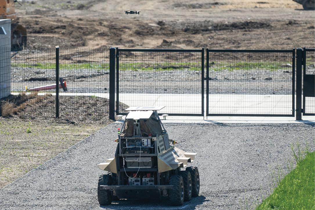 A small drone takes off from the top of a six-wheeled terrain robot.