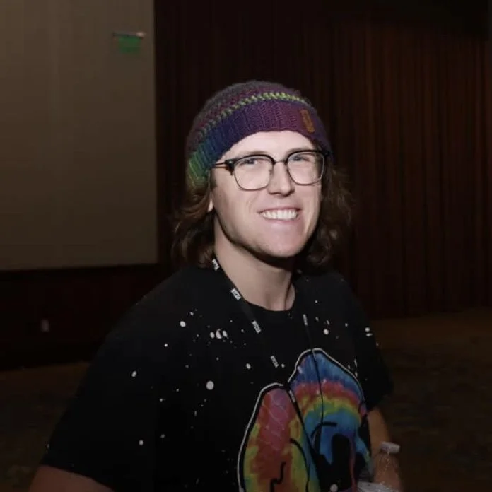 A young man with glasses, long brown hair, wearing a colorful beanie and a black T-shirt with a rainbow-colored design, smiling at the camera indoors.