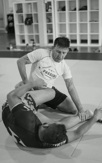 Two men practicing Brazilian Jiu-Jitsu on a mat, one applying a choke hold on the other, with storage shelves in the background.