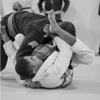 Two men practicing Brazilian Jiu-Jitsu on a mat, one in a white gi and the other in a black gi, engaged in a grappling maneuver.