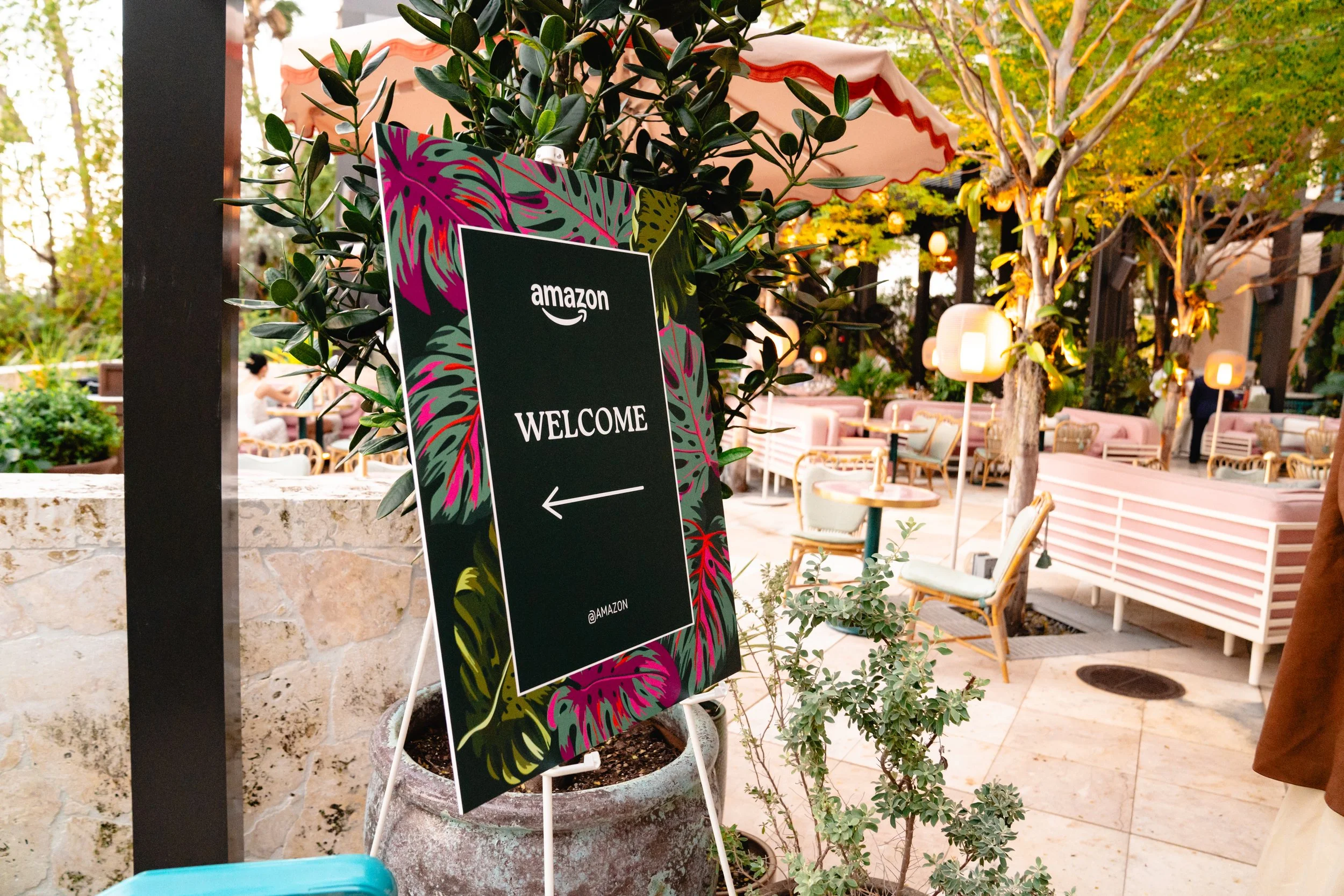 Outdoor restaurant or café seating area with pink and white furniture, trees, and hanging lamps. A black sign with a colorful tropical leaf border reads 'Amazon Welcome' with an arrow pointing left.