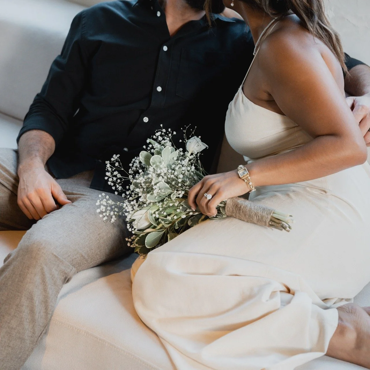 A couple sitting close together; the woman holds a bouquet of white flowers and wears a ring and bracelet.