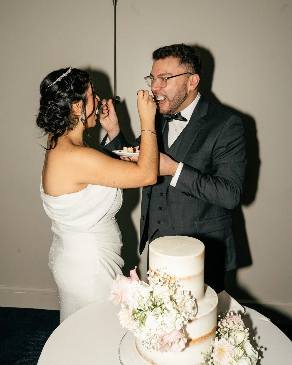Bride and groom in wedding attire feeding each other wedding cake, with a decorated cake on the table.