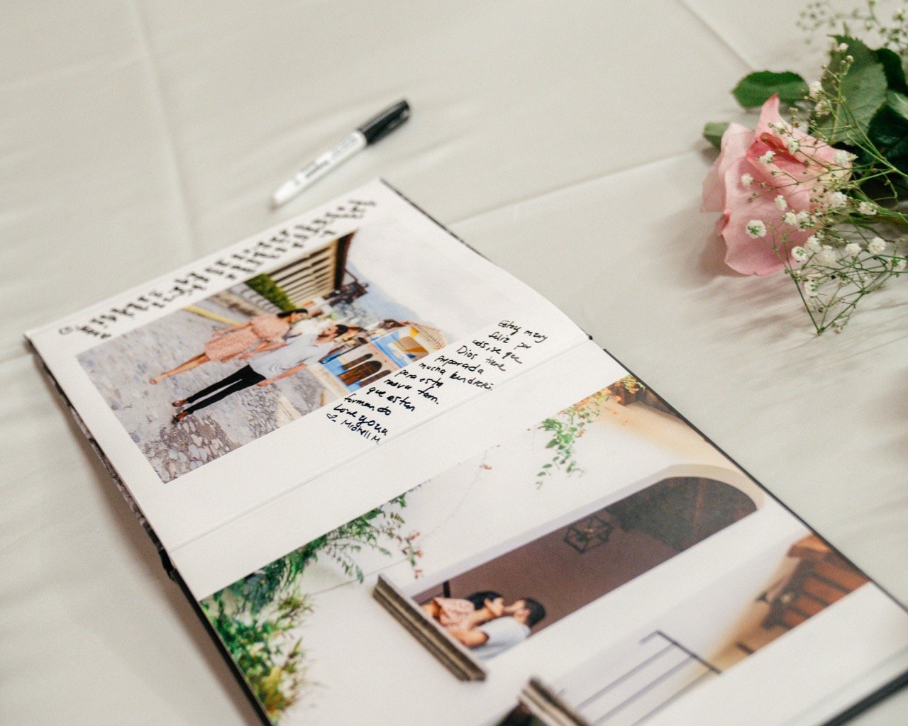 Open photo album with personal pictures and handwritten notes on white surface, accompanied by pink and white flowers nearby.