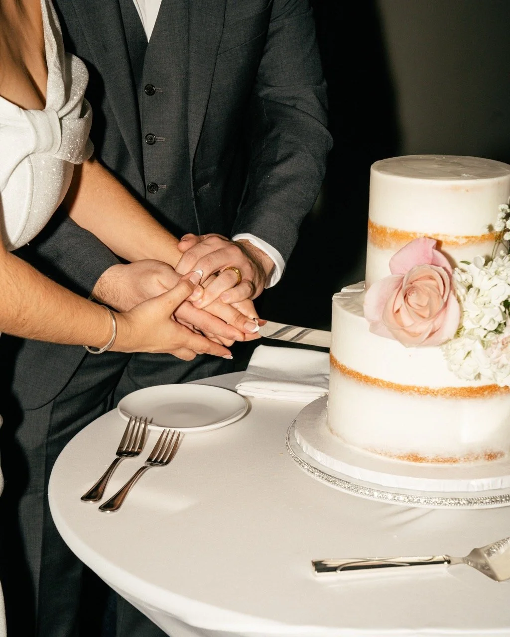 A couple getting married, holding hands with a wedding ring, next to a wedding cake decorated with flowers.