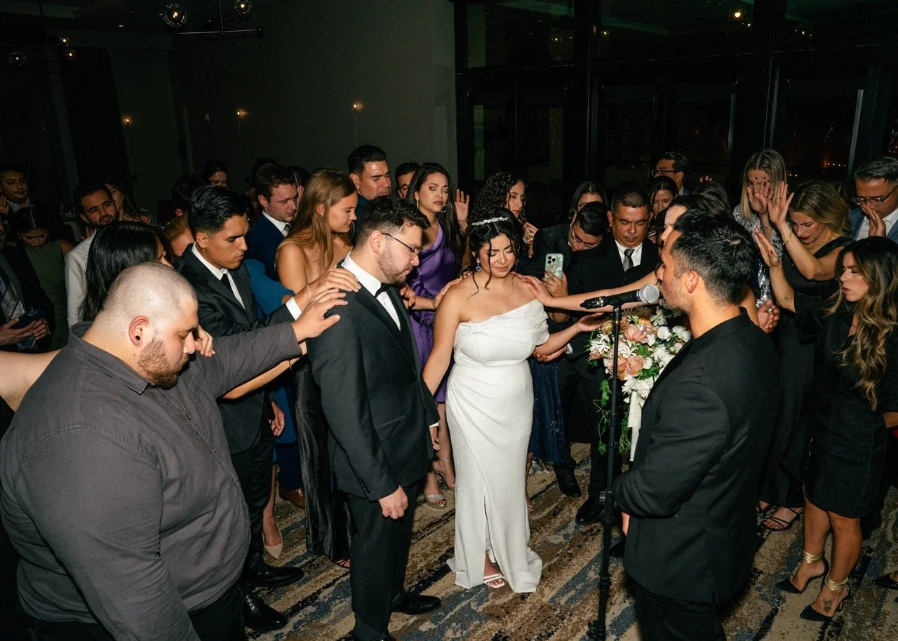 People gathered in a circle at a wedding, with the bride and groom at the center, participating in a prayer or blessing.