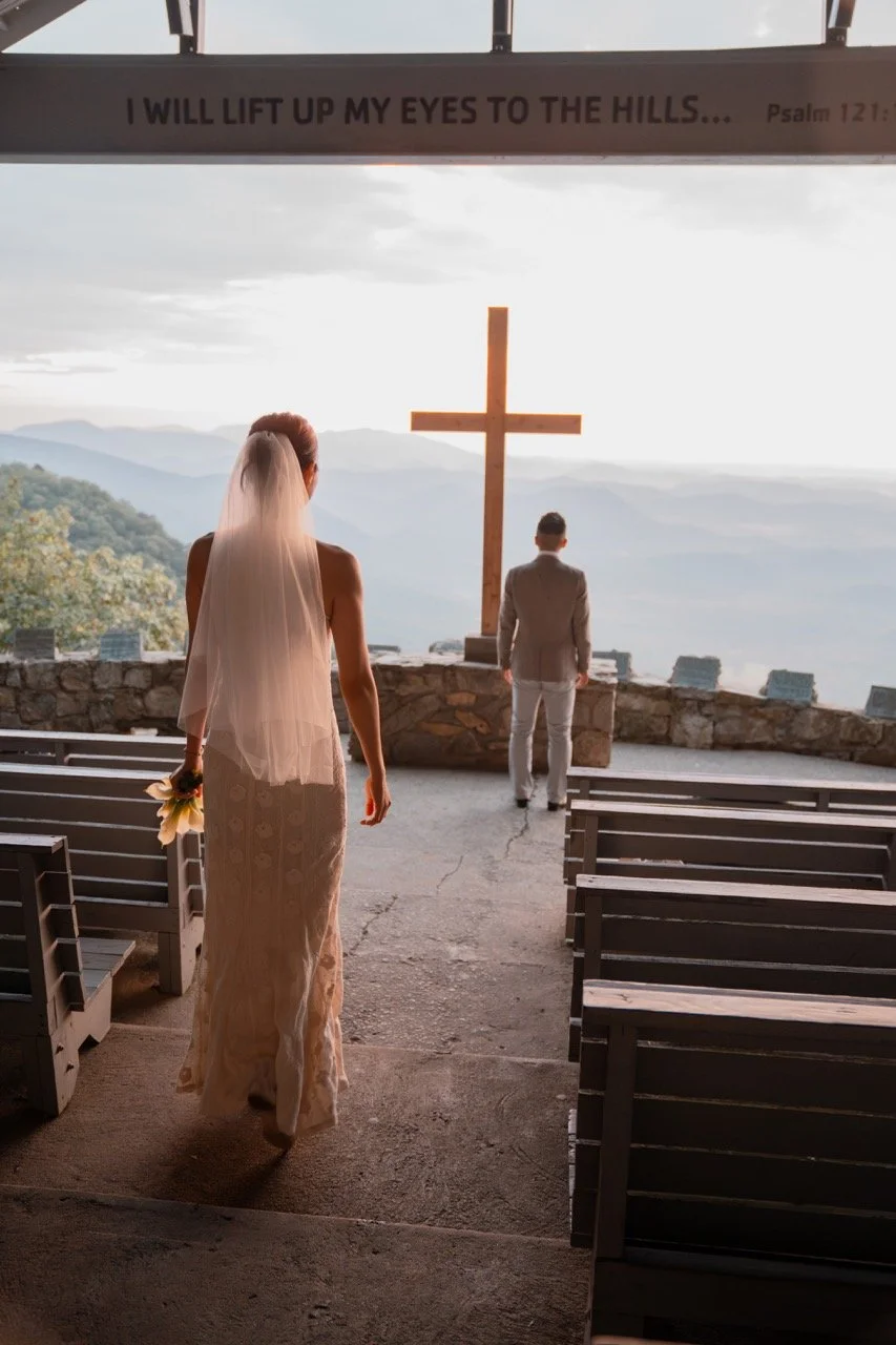 A bride walking down the aisle toward the altar with a cross and mountains in the background, holding a bouquet of flowers, inside a church or chapel.