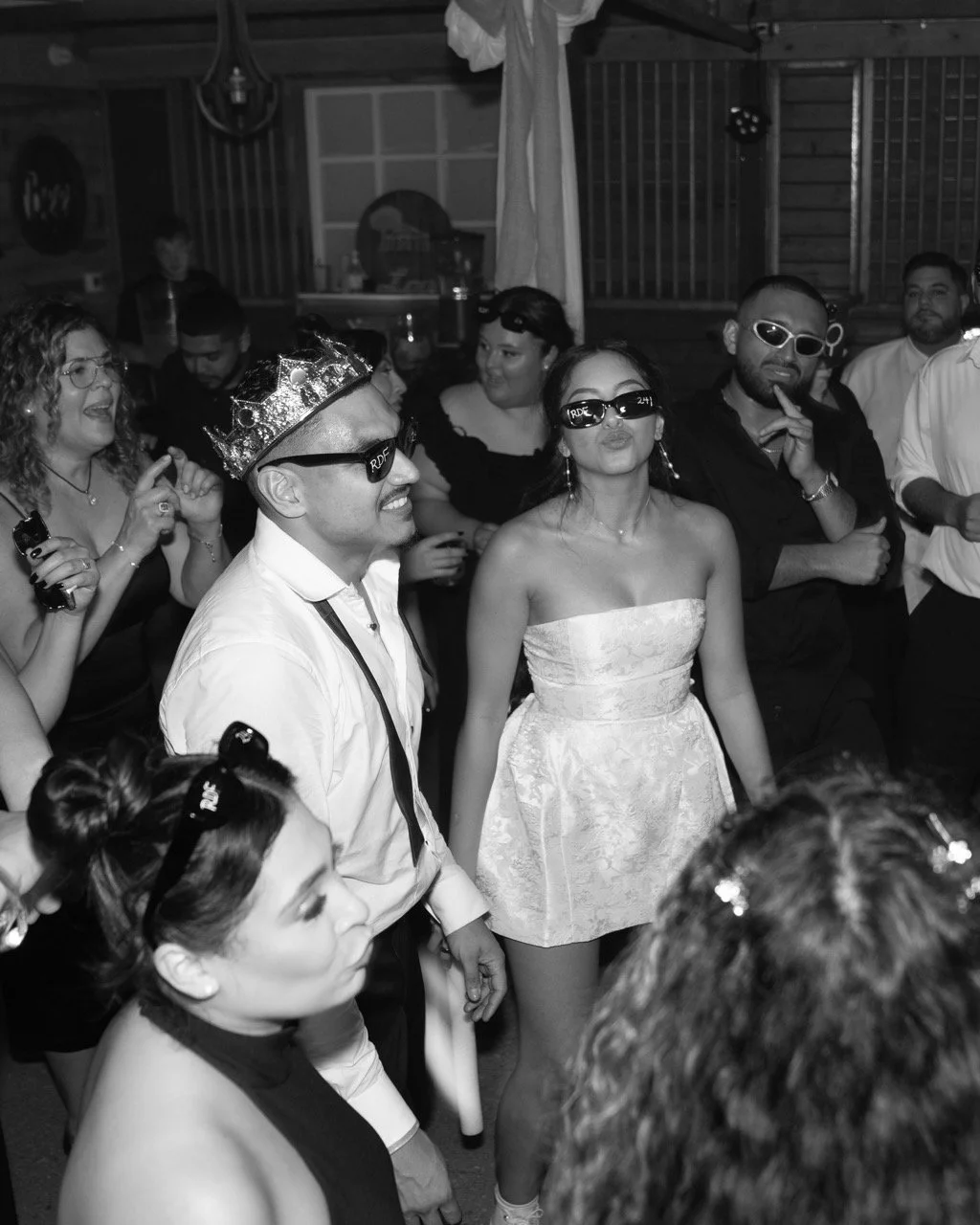 People celebrating at a party, some wearing sunglasses, and one person wearing a crown, all smiling and enjoying the moment in a lively setting.