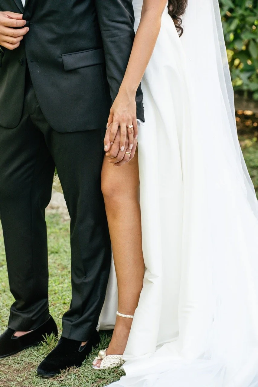 Close-up of a couple holding hands, dressed in wedding attire, outdoors with greenery in the background.