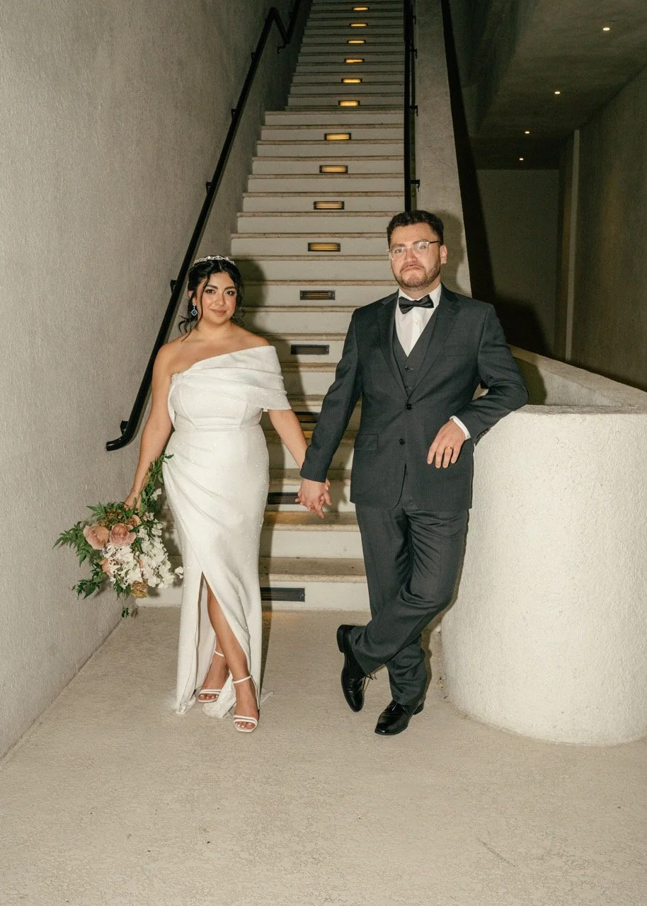 Bride and groom holding hands in wedding attire, standing in front of a staircase at an indoor venue.