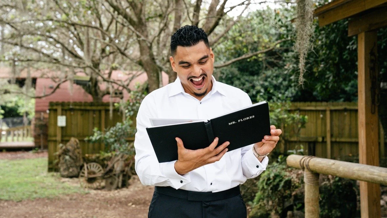 A man in a white dress shirt and black pants is smiling and looking at a black photo album labeled 'Mr. Flores' while standing outdoors in a backyard with trees and a wooden fence.