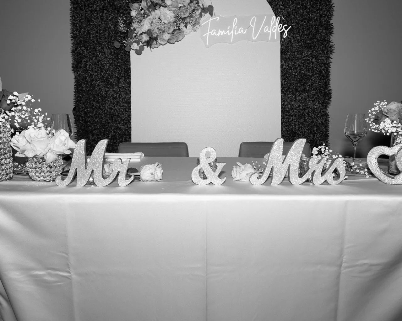 Decorative table setup for a wedding reception with floral arrangements, glassware, and large glittery "Mr" and "Mrs" signs, and a backdrop with a sign that says "Familia Valdes".