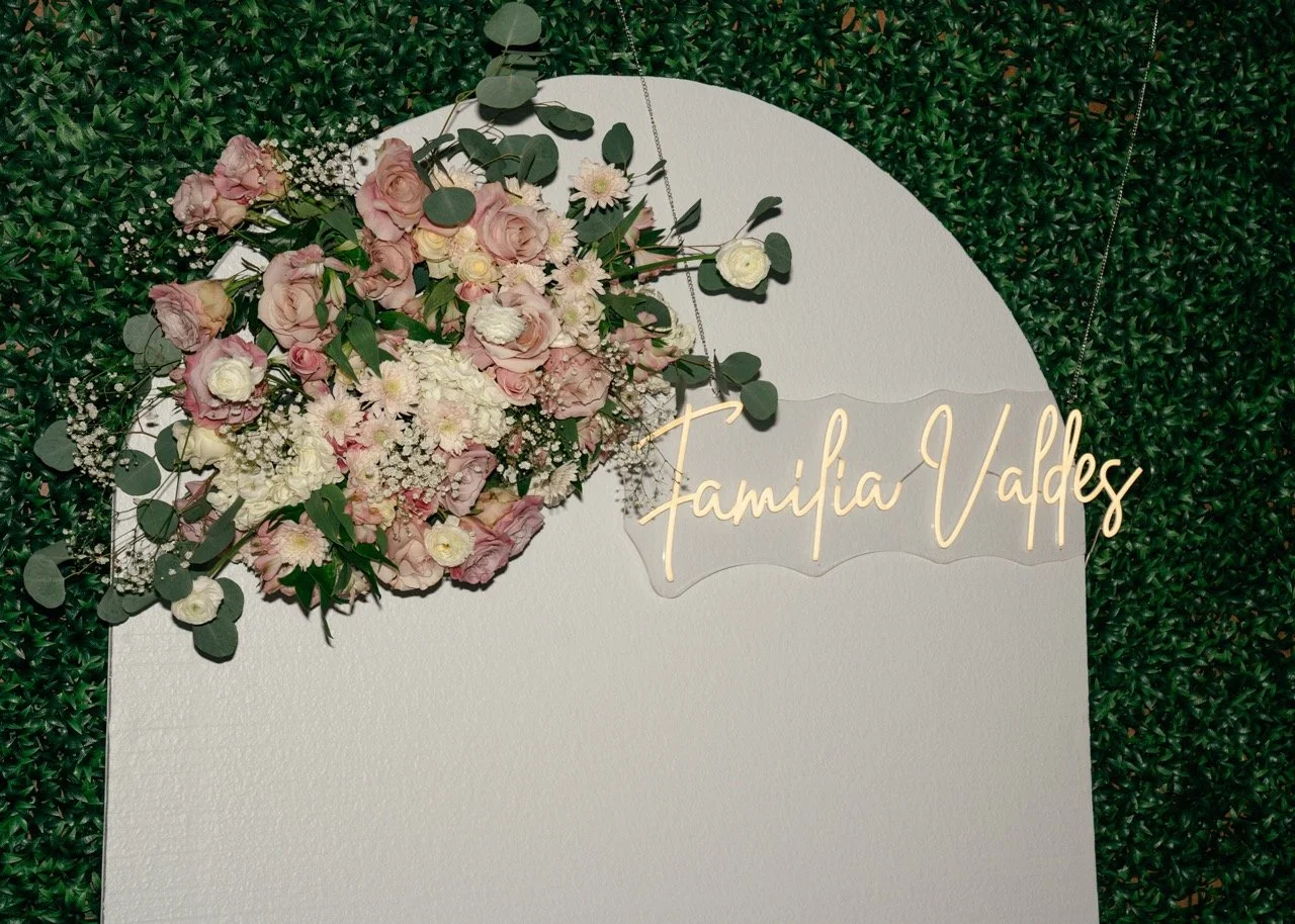 A decorative display with pink and white flowers and green foliage, with a neon sign reading 'Familia Valdes' on a white background, bordered by green leaves.