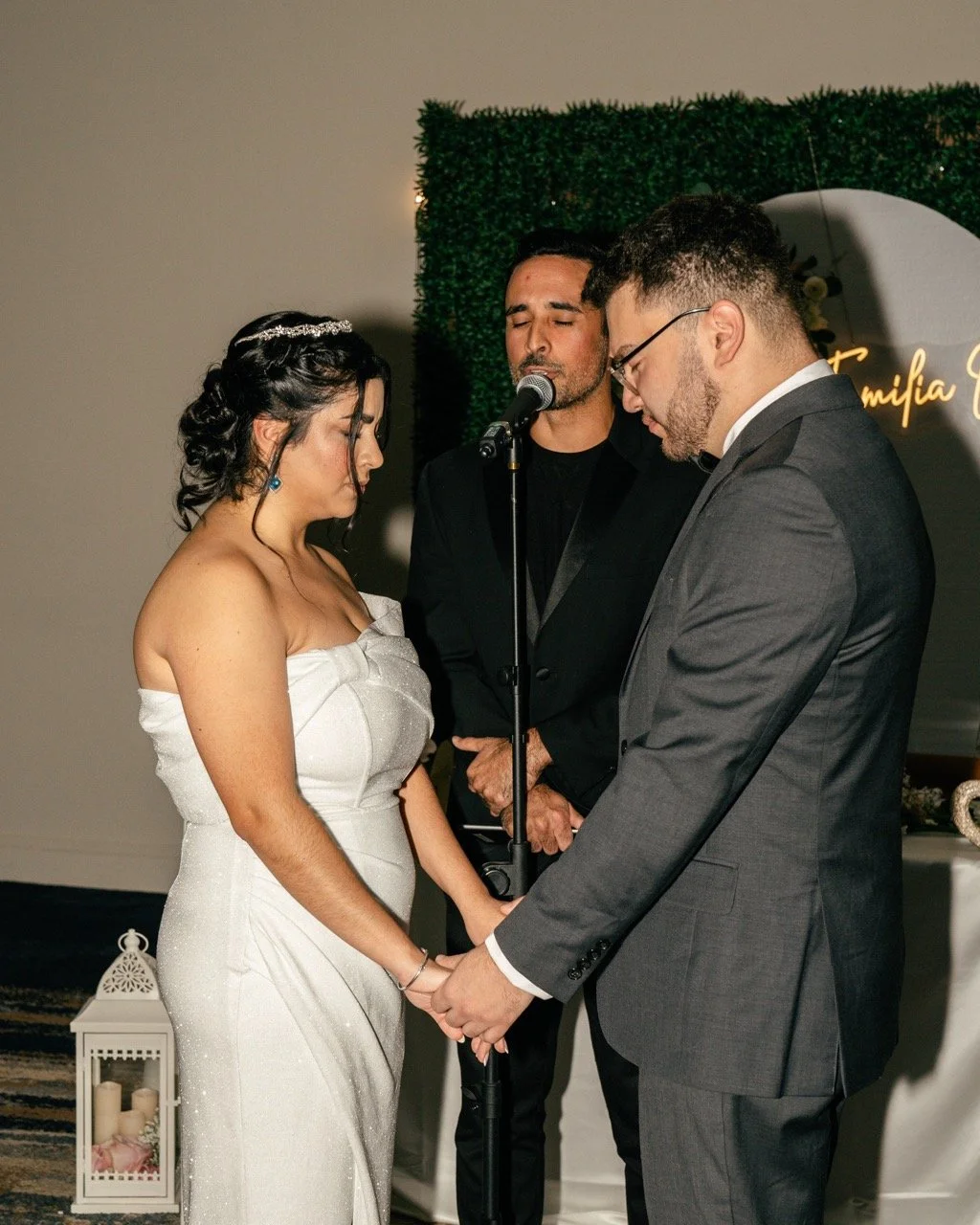 A bride and groom holding hands and praying during their wedding ceremony, with an officiant standing behind them and two candles in a lantern nearby.