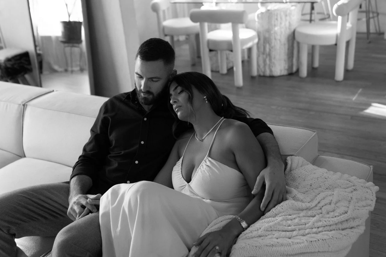 A black and white photo of a man and woman sitting together on a couch, with the man holding her close and the woman leaning her head on his shoulder in an indoor setting.