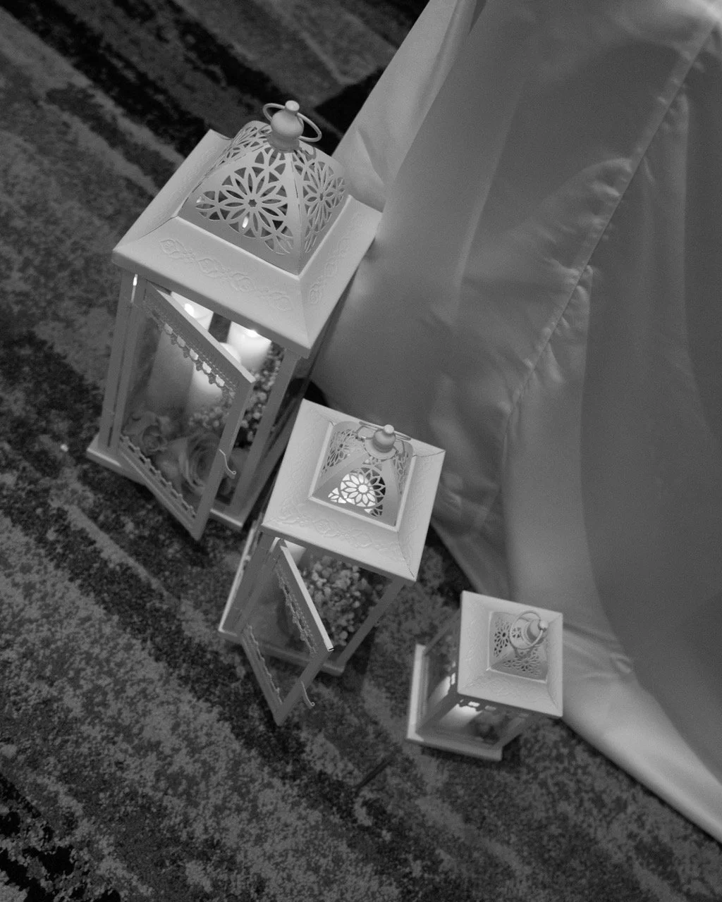 Four white decorative lanterns with intricate cut-out patterns on a patterned carpeted floor, arranged in a cluster near a white fabric backdrop.