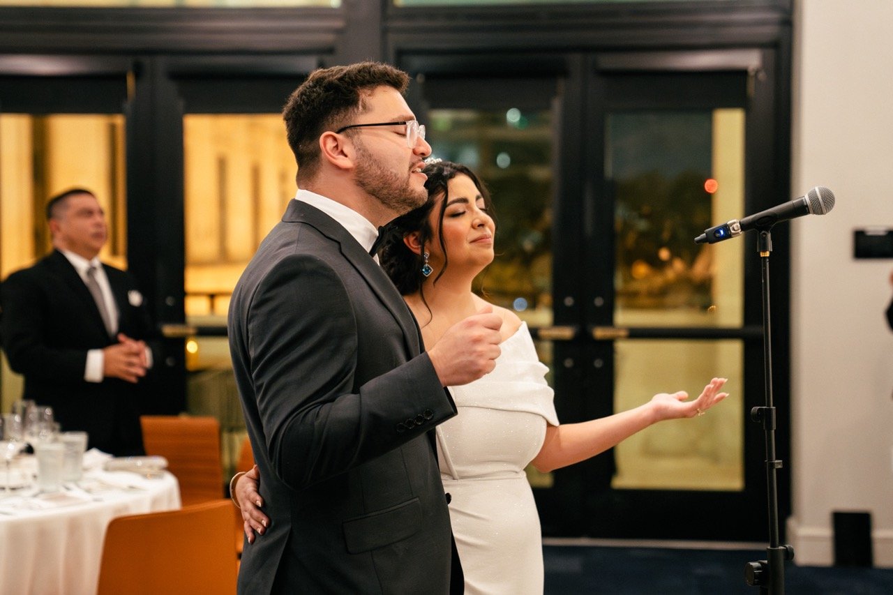 A couple dressed in formal attire, with the man in a tuxedo, appear to be giving a speech or toast at a wedding reception, with a microphone in front of them and guests in the background.