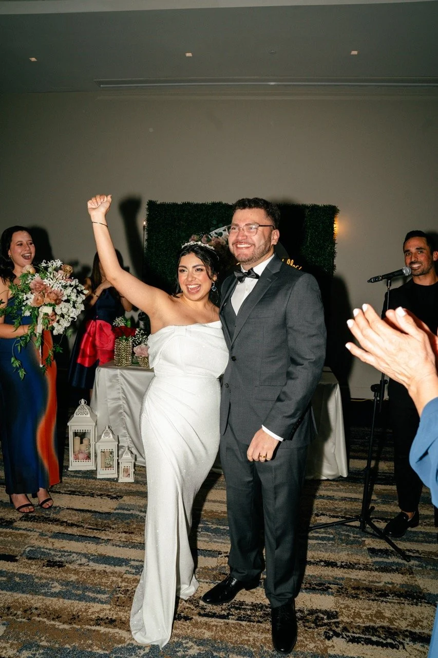 Bride and groom celebrating at their wedding reception, with the bride raising her hand in excitement and the groom smiling.