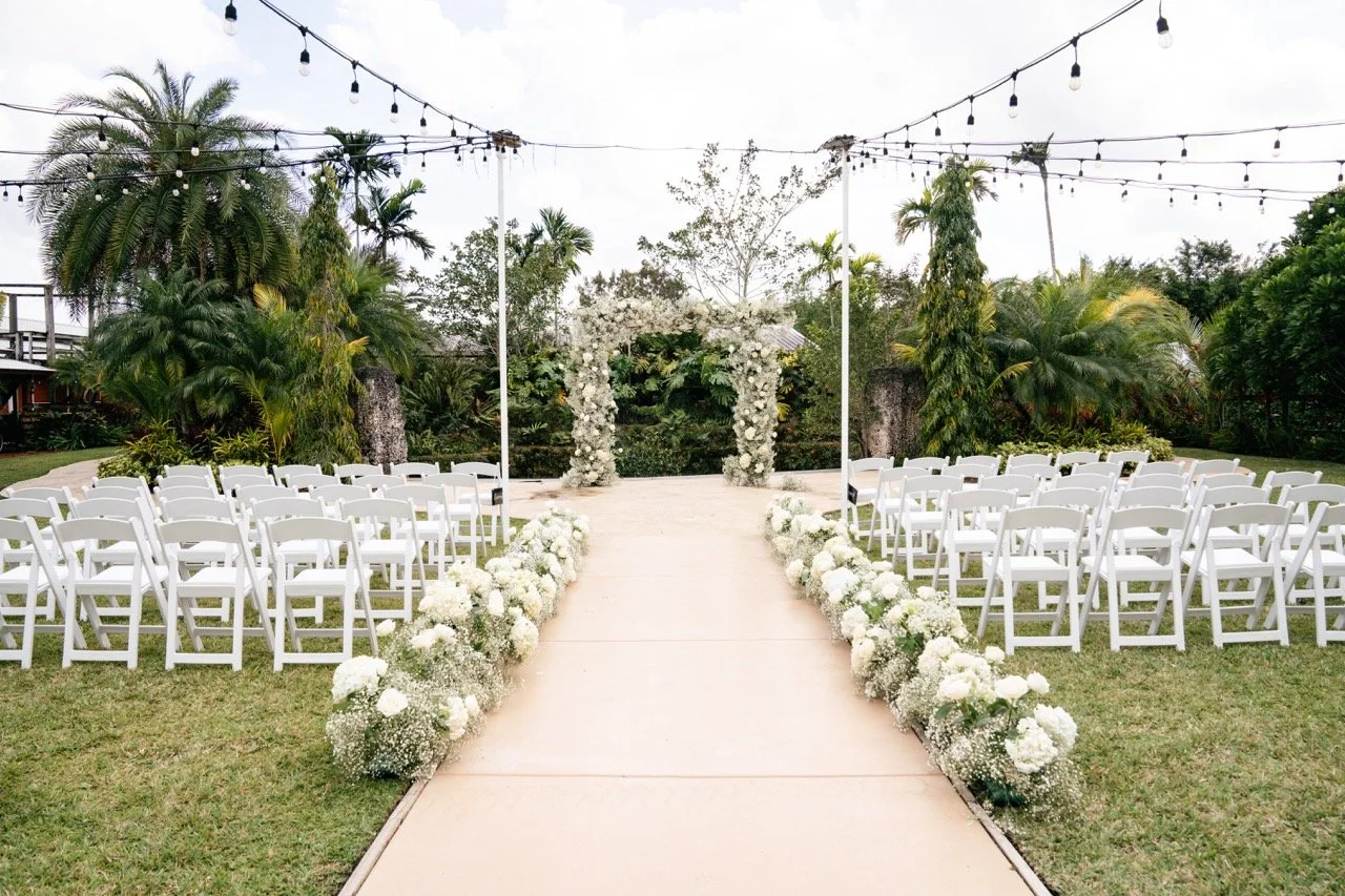 Outdoor wedding ceremony setup with white chairs on either side of a decorated aisle, floral arch at the end of the aisle, surrounded by lush green trees and string lights overhead.