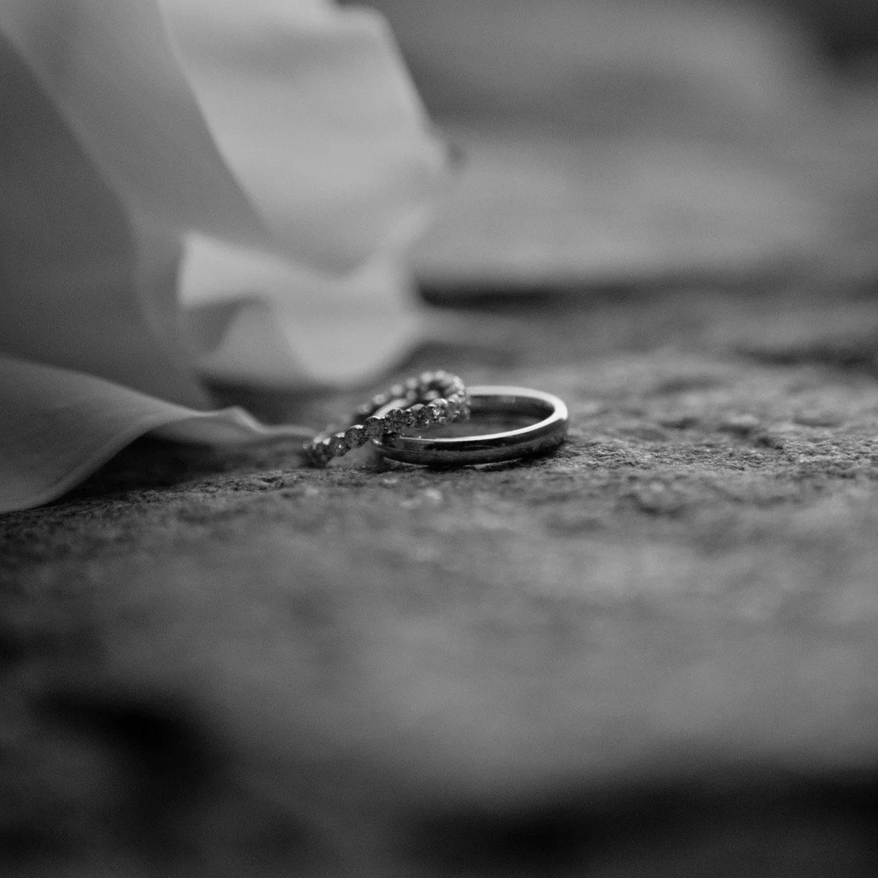 Close-up of wedding rings and a bracelet on a textured surface, with a blurred cloth or paper in the background.