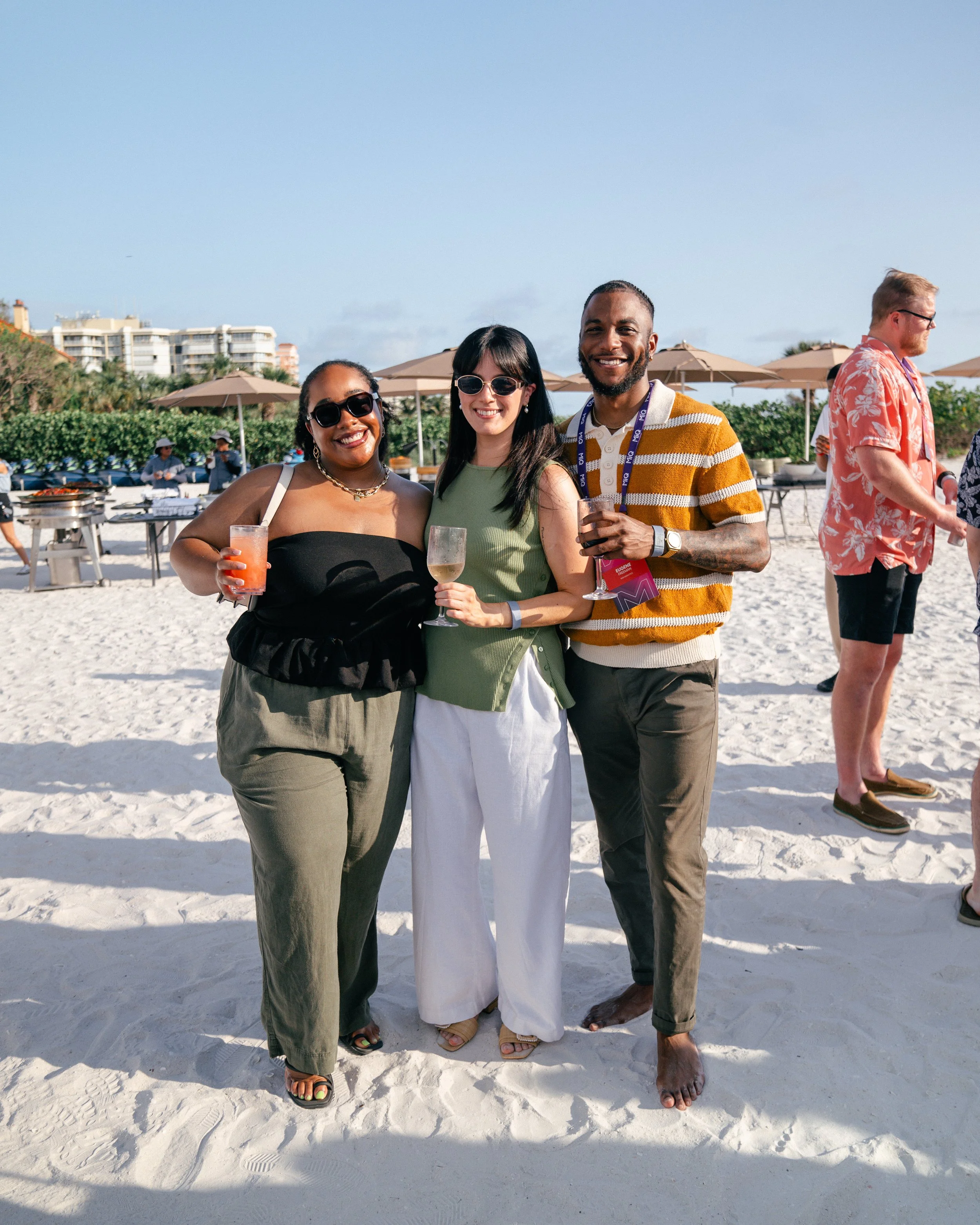 Three friends are smiling and standing together on a sandy beach, each holding a drink, during daytime. In the background, other beachgoers, umbrellas, and buildings are visible.