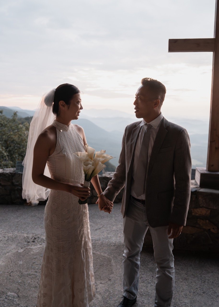 A bride and groom holding hands and facing each other on a mountain overlook at sunset, with a scenic view of mountains in the background.