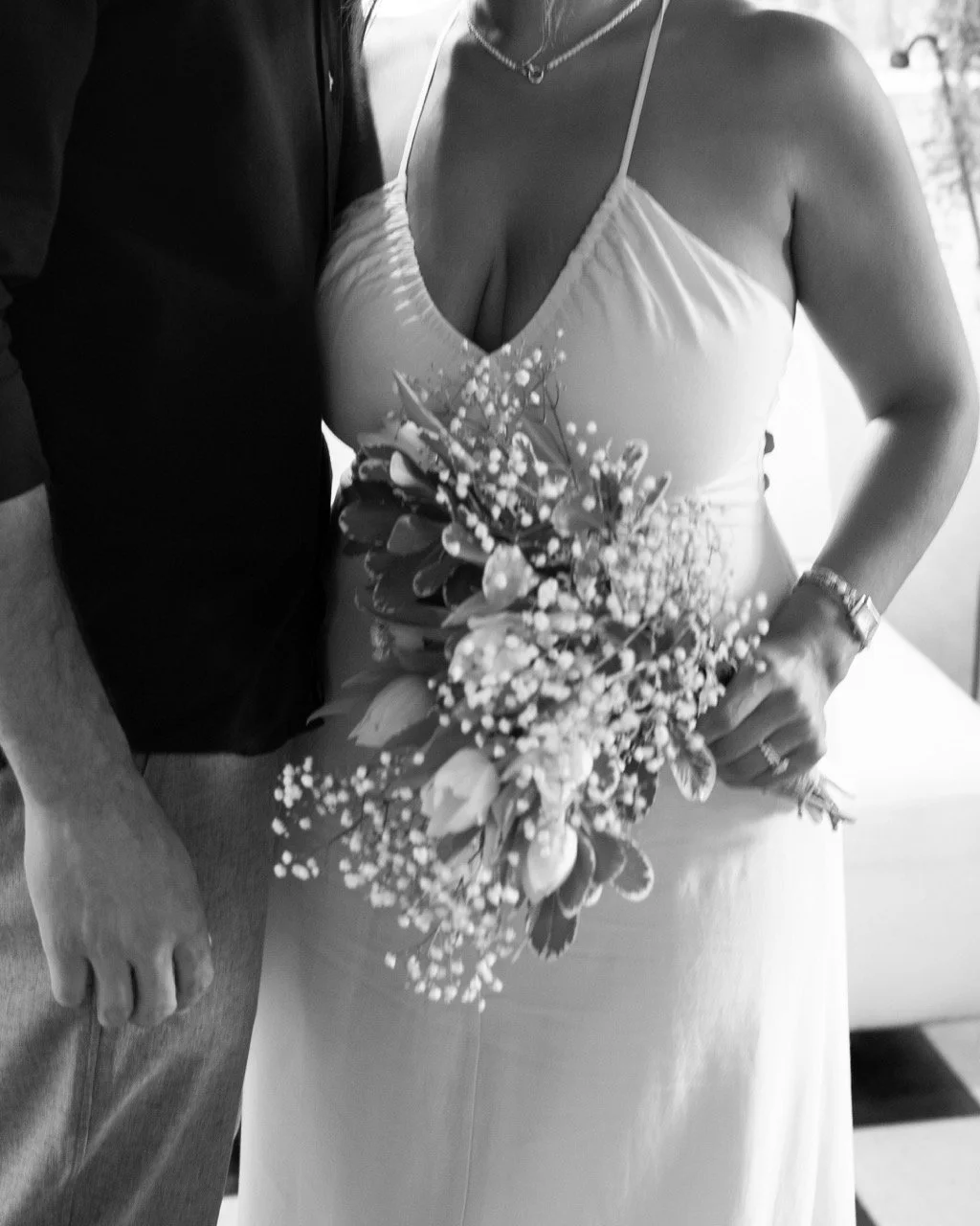 A woman in a wedding dress holding a bouquet of flowers, standing next to a man in a dark shirt. The woman is wearing a wedding gown with spaghetti straps and a necklace. The photo is in black and white.