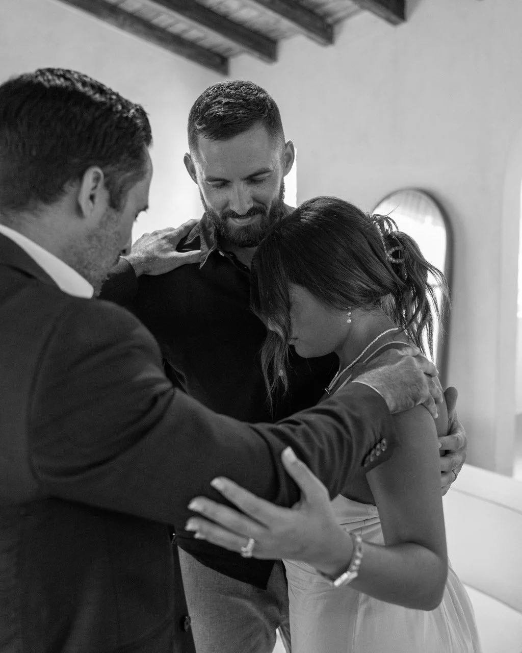 Three people engage in a prayer or moment of reflection, with heads bowed and hands on each other's shoulders inside a room.