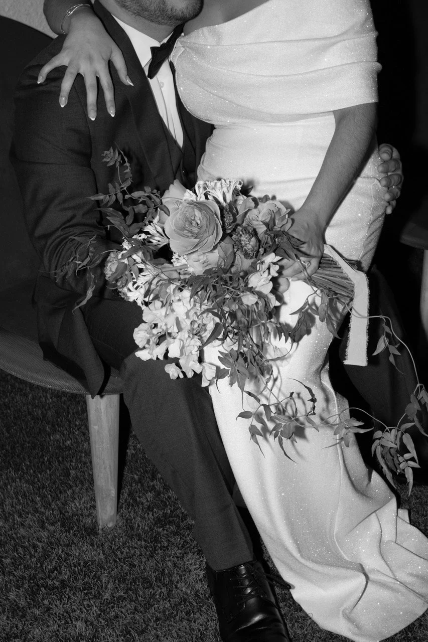 A person in a tuxedo holding a wedding bouquet, sitting with a person in a wedding gown leaning on him.