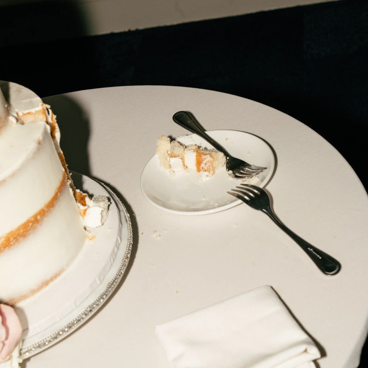 A few bites of cake on a small plate with a spoon and fork, placed on a white table next to a larger cake.
