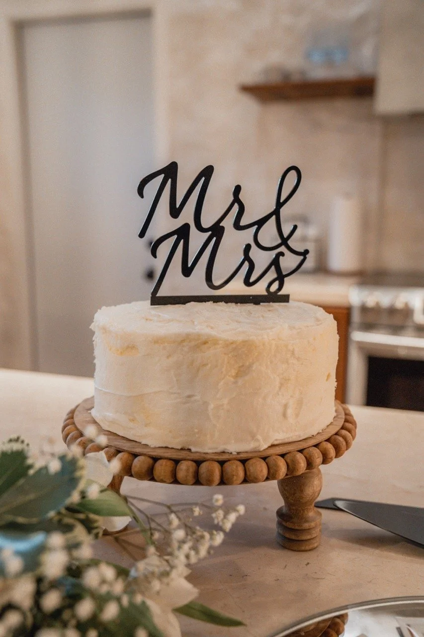 A white frosted cake on a wooden cake stand with a "Mr & Mrs" topper, placed on a table with flowers and a smartphone nearby in a cozy kitchen setting.