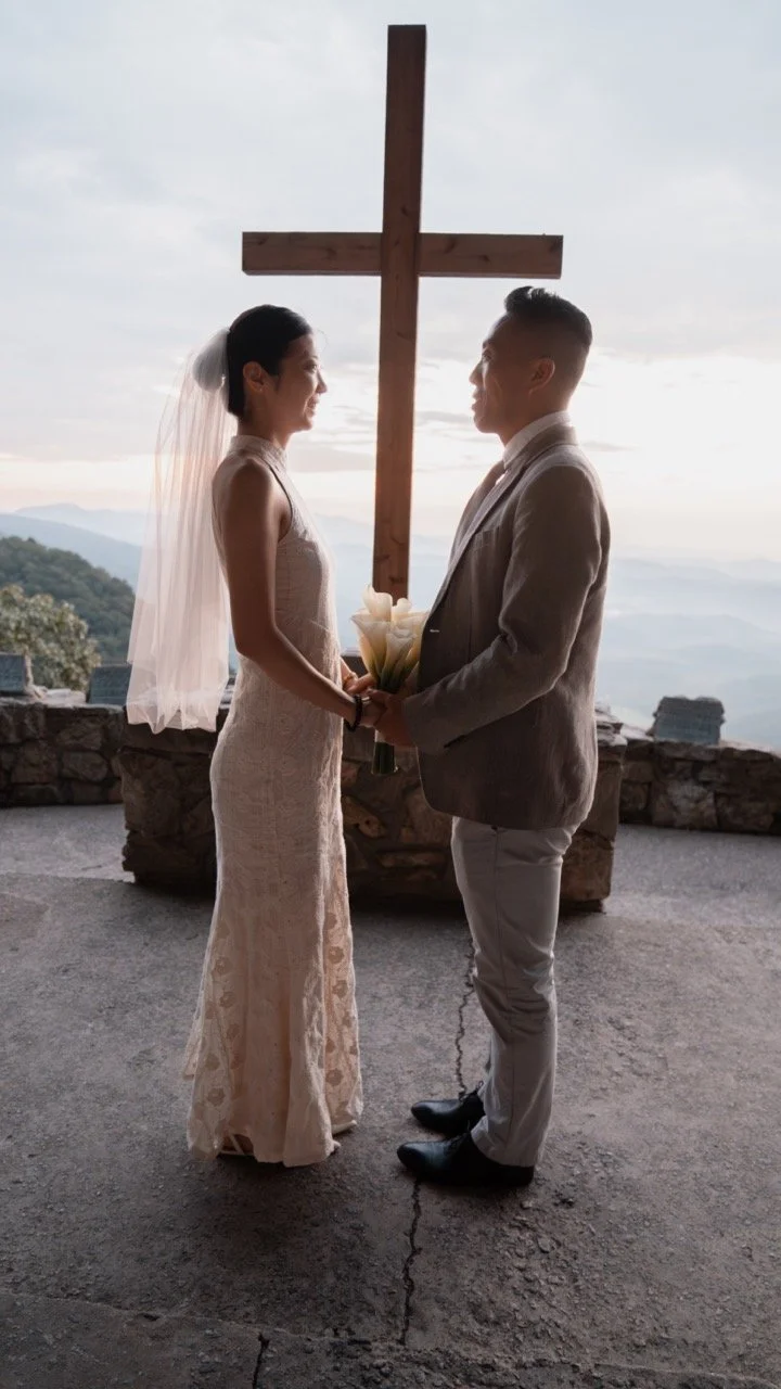 A bride and groom standing face to face, holding a bouquet, with a large wooden cross behind them, overlooking a mountainous landscape during a wedding ceremony.