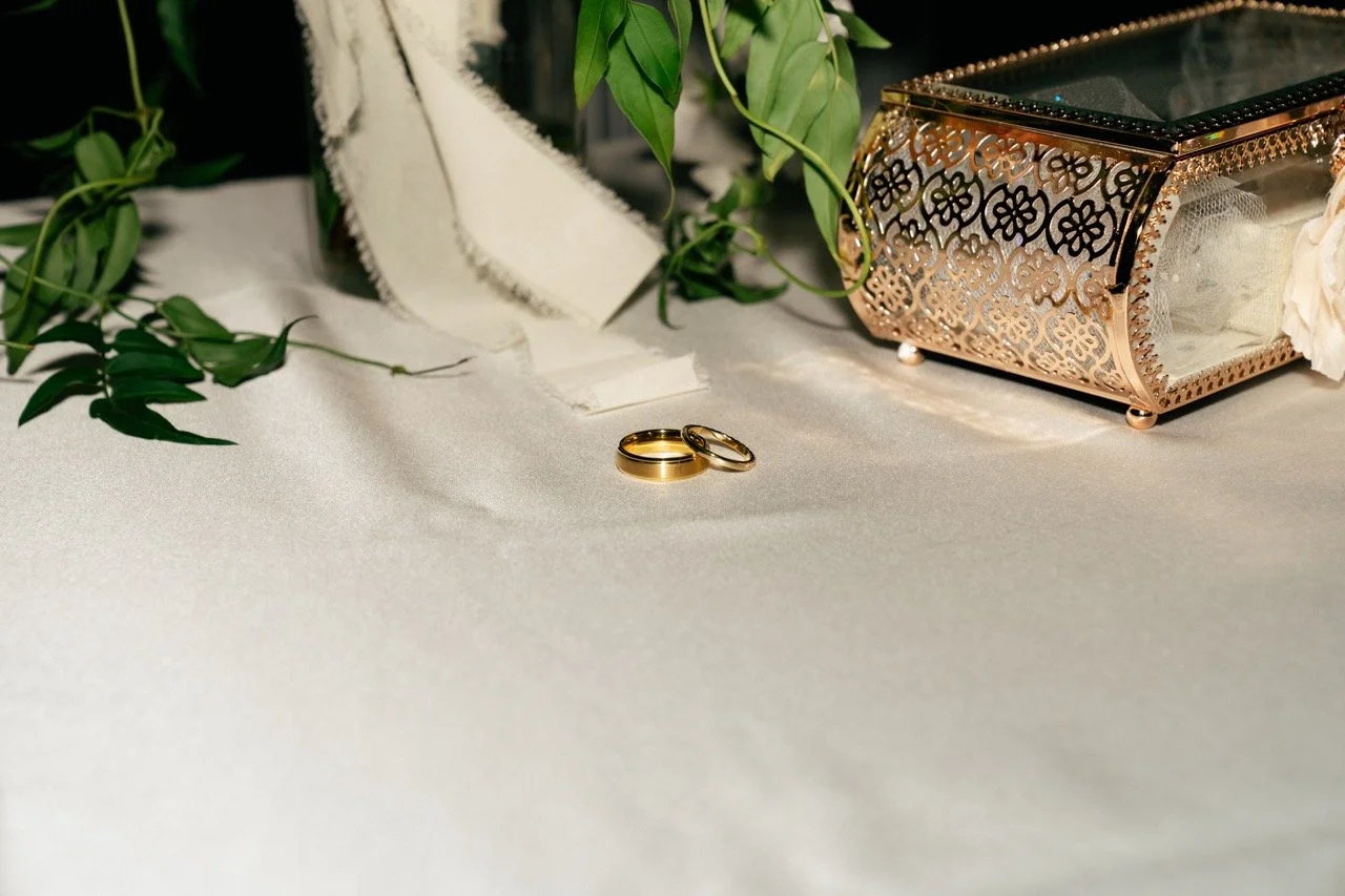 A pair of gold rings resting on a white fabric surface, surrounded by a decorative jewelry box, green leaves, and a fabric cloth.