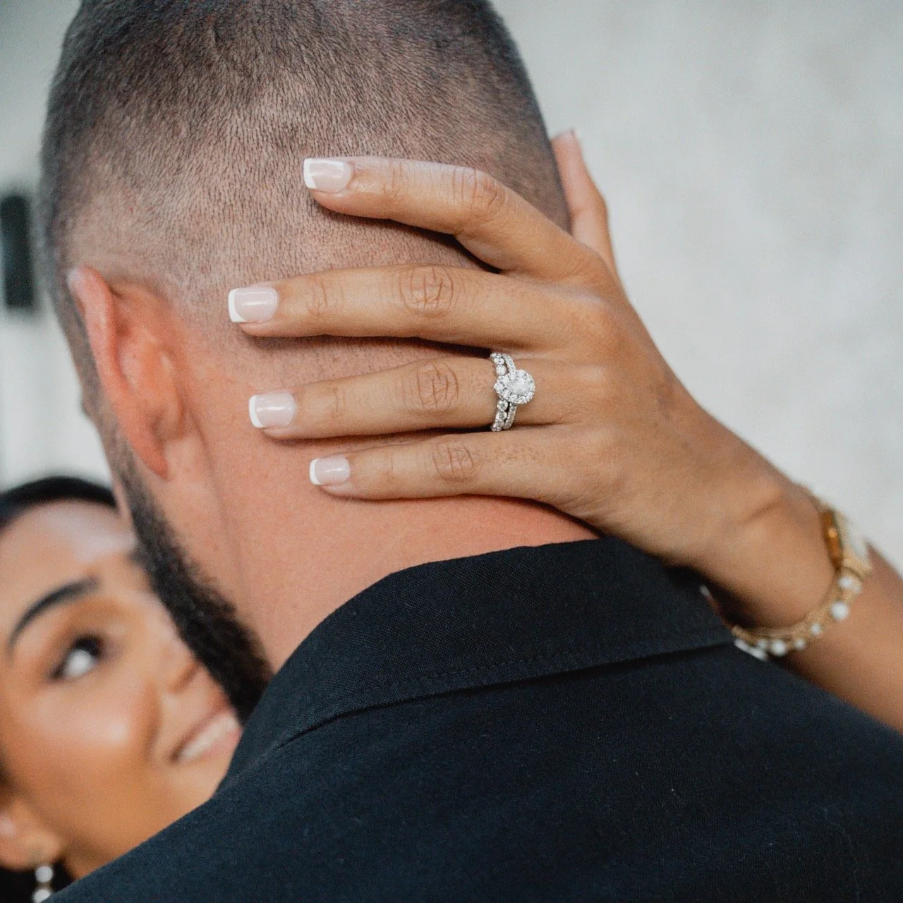Close-up of a woman’s hand with a wedding ring on her finger, resting on a man's neck during a wedding kiss. The woman has well-manicured nails, a bracelet, and a ring with multiple diamonds. A bride with makeup and earrings is visible out of focus i