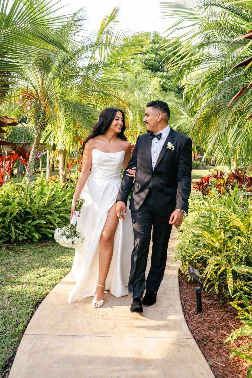 A bride and groom walking arm-in-arm on a garden pathway surrounded by lush tropical greenery, with the bride holding a bouquet and wearing a white strapless gown, and the groom in a black tuxedo with a bow tie, both smiling at each other.