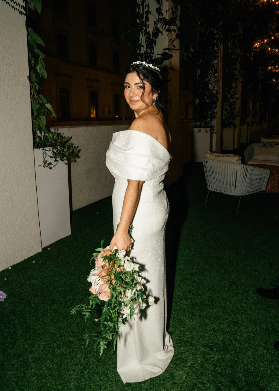 A woman in a white off-the-shoulder gown holding a bouquet of pink and white flowers, standing on a green outdoor area at night with buildings and string lights in the background.