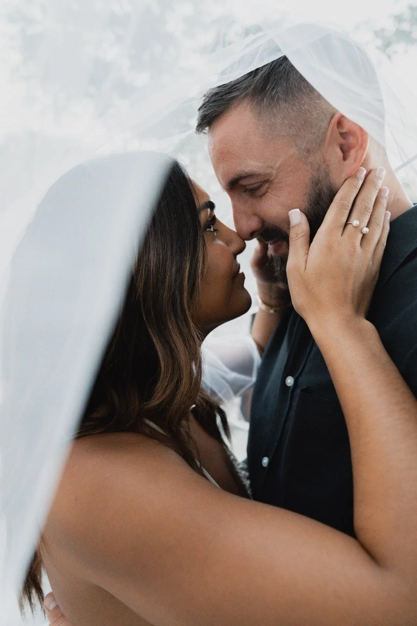 A bride and groom face to face, close up, inside a wedding veil, sharing an intimate moment during their wedding.