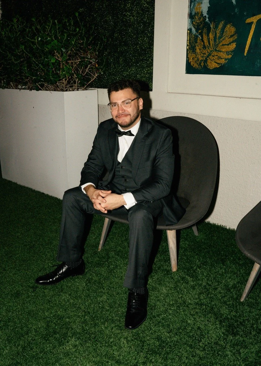 A man dressed in a black tuxedo with a bow tie, glasses, and a beard sitting on a black chair outdoors on green artificial grass, with a white wall and potted plants in the background.