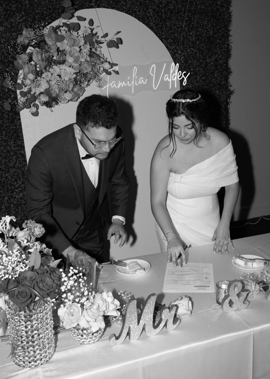 Black and white photo of a bride and groom signing a marriage certificate at a wedding reception table decorated with flowers and a "Mr & Mrs" sign.